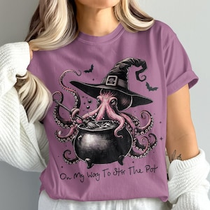 May include: A purple t-shirt featuring a whimsical illustration of an octopus wearing a witch's hat, stirring a cauldron. The text "On My Way To Stir The Pot" is below the image, with bats in the background.