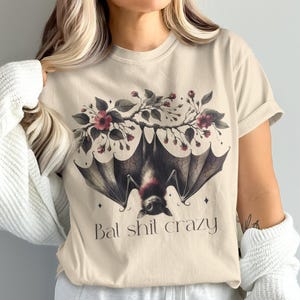 Puede incluir: Camiseta beige con un diseño de murciélago, elementos florales y el texto "Bal shit crazy". El murciélago está boca abajo con las alas extendidas, complementado con flores rojo oscuro y hojas verdes.