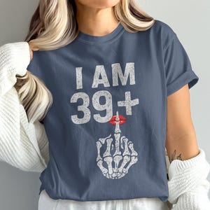 Op de afbeelding: Een marineblauw t-shirt met de witte tekst "I AM 39+", een rode lippenstiftkus en een skelet hand die de middelvinger opsteekt. Het shirt heeft een casual stijl.
