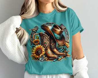 Puede incluir: Camiseta turquesa con un diseño de temática western. El diseño incluye un sombrero de vaquero marrón con una banda turquesa, botas de vaquero marrones, girasoles y una herradura. Las botas y el sombrero están delante de la herradura.