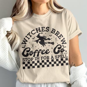 May include: Beige t-shirt with a black graphic that reads "Witches Brew Coffee Co. Stop in for a Spell." The design includes a witch on a broom, bats, and a checkered pattern.