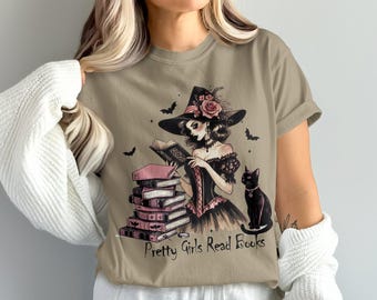 May include: Tan t-shirt featuring a witch reading a book, with a black cat and bats. The witch wears a black hat with roses and a corset. A stack of pink books is next to her. The text "Pretty Girls Read Books" is at the bottom.