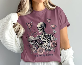May include: Mauve t-shirt with a skeleton reading a book, surrounded by flowers and butterflies. The text "Book Are My Therapy" is printed below the illustration. The design incorporates pink and cream colours.