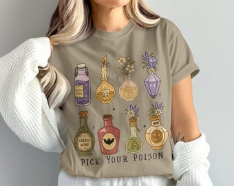 May include: A tan t-shirt featuring a collection of illustrated potion bottles in various shapes and colors. The bottles are filled with liquids and adorned with flowers, candles, and crystals. The text "PICK YOUR POISON" is printed below the bottles.