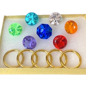 May include: A collection of colorful, faceted gemstones in various shapes and sizes, including round and octagonal, presented in a gold box. Accompanying the gemstones are six gold-colored metal rings.