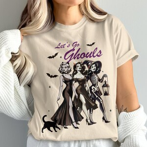 May include: Beige t-shirt with a Halloween-themed graphic. The design features three women in dark dresses, bats, and the text "Let's Go Ghouls". A black cat is also included in the illustration.