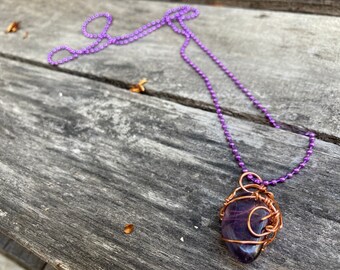 Op de afbeelding: Een paarse amethiststeen gewikkeld in koperdraad, hangend aan een paarse kralenketting.