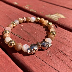 May include: A beaded bracelet with gold accents and two silver skull charms. The beads are a mix of light brown, pink, and white.