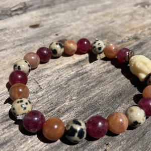 Pode incluir: Um bracelete de contas com um amuleto de Buda branco. O bracelete apresenta contas vermelhas, marrons e brancas com manchas pretas.