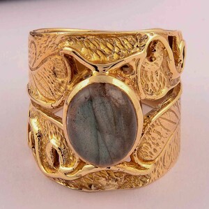 Puede incluir: Un anillo ancho de oro con una piedra preciosa ovalada de color gris verdoso. El anillo tiene un diseño ornamentado y texturizado con patrones en forma de hojas. La piedra preciosa está engastada en el centro del anillo.
