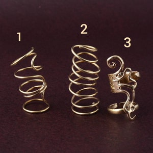 Puede incluir: Tres anillos dorados con diseños únicos en espiral y abstractos. Los anillos se muestran sobre una superficie marrón oscuro. El anillo 1 presenta un diseño en espiral, el anillo 2 tiene una espiral apretada y el anillo 3 tiene un diseño adornado y abierto.