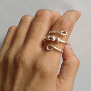 Puede incluir: Un anillo plateado con un diseño en espiral, con tres pequeñas cuentas redondas y extremos en bucle. El anillo se lleva en un dedo, mostrando su estilo intrincado y minimalista. La joya está hecha de un metal brillante.