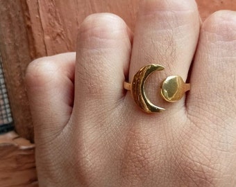 May include: Gold-colored ring featuring a crescent moon and a circular accent. The ring has a textured surface and is worn on a finger. The ring's design is simple and elegant, suitable for everyday wear.