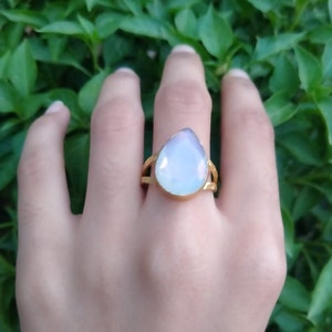 Puede incluir: Un anillo dorado con una gran piedra preciosa azul pálido en forma de lágrima. El anillo tiene un diseño de doble lazo en la banda. El anillo se muestra en una mano, sobre un fondo de follaje verde.