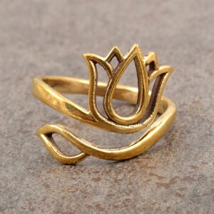 Puede incluir: Un anillo dorado con un diseño de flor de tulipán estilizada. El anillo se envuelve alrededor del dedo con una parte superior floral calada. El anillo está hecho de un material metálico brillante y se encuentra sobre una superficie de tono neutro.