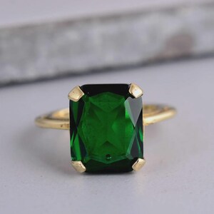 Puede incluir: Un anillo de oro con una gran piedra preciosa rectangular de color verde esmeralda facetada, montada en un engaste de cuatro garras. La banda es fina y de oro metálico, proporcionando un marco sencillo para la piedra verde intenso central.