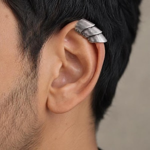 Puede incluir: Un ear cuff de tono plateado con un diseño segmentado y superpuesto que recuerda a una armadura articulada. La pieza de metal tiene un acabado mate y desgastado con pequeñas hendiduras circulares en el lateral, ajustada al cartílago superior.