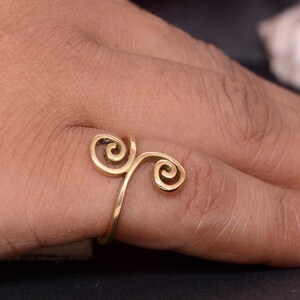 Puede incluir: Un anillo dorado con un diseño de espiral único. El anillo presenta dos espirales arremolinadas conectadas por una banda delgada, creando un aspecto elegante y artístico. El anillo se muestra en el dedo de una persona.