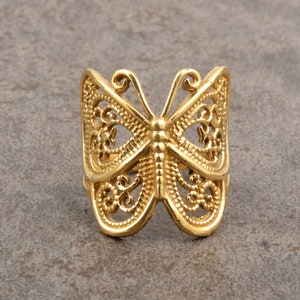 Puede incluir: Un anillo de mariposa dorado. El anillo presenta un diseño calado con detalles intrincados, mostrando las alas y el cuerpo de la mariposa. El anillo está hecho de un material metálico brillante.