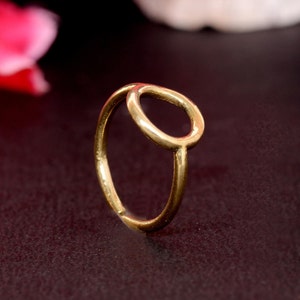 Puede incluir: Un anillo dorado con un diseño ovalado abierto. El anillo está hecho de un metal brillante y tiene un estilo simple y elegante. El anillo se muestra sobre un fondo oscuro con una flor rosa borrosa.