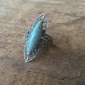 Puede incluir: Un anillo con una piedra de color azul claro en forma de marquesa con vetas oscuras, engastada en una banda de metal plateado con intrincados detalles de filigrana calada que se extienden hacia afuera en un patrón de hojas.