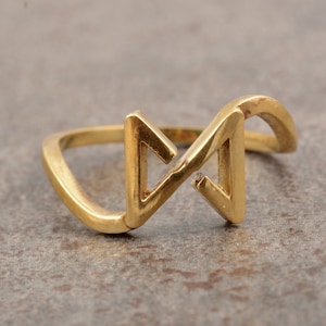 Puede incluir: Un anillo dorado con un diseño geométrico. El anillo presenta una forma estilizada de la letra "N" con una pequeña piedra de acento transparente. La banda es lisa y redondeada, y el diseño general es moderno y minimalista.