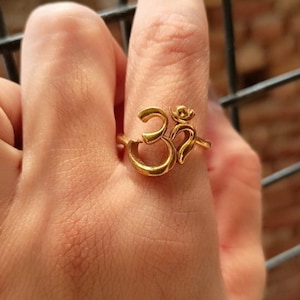 Puede incluir: Un anillo dorado con el símbolo Om. El anillo se lleva en un dedo, mostrando el intrincado diseño y el cálido tono metálico de la joya. El símbolo Om es un icono espiritual.