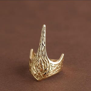 Puede incluir: Un anillo de metal en tono dorado que presenta una extensión vertical alargada y puntiaguda que se eleva desde el centro de la banda. La superficie del anillo está decorada con intrincados patrones de remolinos en relieve que cubren toda la estructura.