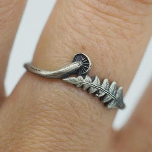 Puede incluir: Un anillo de metal plateado con un diseño detallado y texturizado de un pequeño sombrero de hongo conectado a una hoja de helecho. El metal tiene un acabado oxidado que resalta las intrincadas ranuras y las formas naturales botánicas.