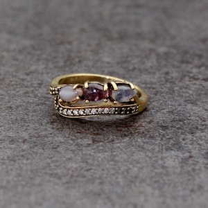 Puede incluir: Un anillo dorado con tres piedras preciosas ovaladas en tonos rosa, morado y gris. El anillo tiene una banda con pequeñas piedras claras y un diseño decorativo. El anillo está sobre un fondo neutro.