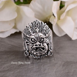 Puede incluir: Anillo plateado con un diseño de rostro estilizado y detallado, con boca abierta y tocado ornamentado. El anillo tiene un diseño llamativo. El texto "Jazzy Ring Store" es visible en la imagen.