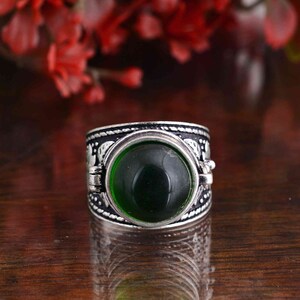 Puede incluir: Un anillo plateado con una piedra grande, redonda y verde oscuro en el centro. La banda presenta un intrincado patrón con pequeñas figuras de elefantes y un borde texturizado. El anillo está sobre un fondo borroso de flores rojas.