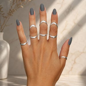 Puede incluir: Un conjunto de seis anillos de tono plateado usados en los dedos. Tres anillos son bandas simples, mientras que tres presentan un diseño de chevrón en forma de V posicionado en las articulaciones superiores. Tienen un acabado metálico pulido.
