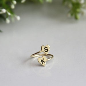 Puede incluir: Anillo dorado con dos colgantes en forma de corazón. Cada colgante tiene una letra negra, "S" en el corazón superior y "K" en el inferior. El anillo está sobre una superficie blanca.