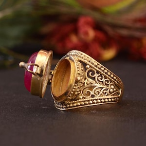 May include: A gold-colored ring with an open compartment and a hinged lid. The lid features a red gemstone. The ring has an ornate, filigree design on the band.
