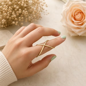 Puede incluir: Un anillo de tono dorado con una banda fina y lisa que se cruza sobre sí misma para formar una X. El anillo se lleva en un dedo, mostrando su diseño abierto y sencillo con un acabado metálico pulido que refleja la luz en su superficie.