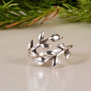 Puede incluir: Un anillo plateado con un diseño de hojas. El anillo presenta múltiples hojas que se envuelven alrededor del dedo. Las hojas tienen una textura detallada y están dispuestas en un patrón natural y orgánico. El anillo está sobre un fondo neutro.