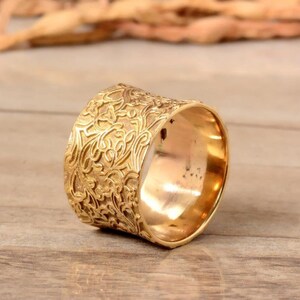 Puede incluir: Un anillo ancho de color dorado con un intrincado diseño floral. La superficie del anillo está texturizada con diseños detallados en relieve. El anillo se muestra sobre una superficie de madera, mostrando su cálido tono metálico. Este anillo es una joya.