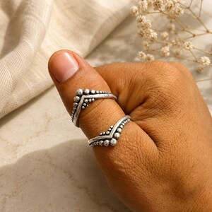 Puede incluir: Dos anillos de tono plateado con un diseño en forma de V o chevron. Cada anillo está adornado con una serie de pequeñas cuentas esféricas en relieve a lo largo del borde exterior, creando un acabado metálico texturizado.