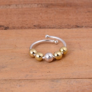 Puede incluir: Un anillo abierto con una banda fina de color plateado y extremos redondeados. El anillo está adornado con cinco pequeñas cuentas lisas dispuestas en la parte frontal, compuestas por cuatro cuentas doradas y una cuenta plateada central.