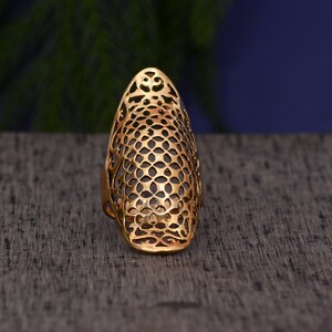 Puede incluir: Un anillo dorado con un intrincado diseño calado. El anillo presenta una forma ovalada alargada con un patrón detallado de círculos interconectados y formas florales. El anillo está sobre un fondo neutro.
