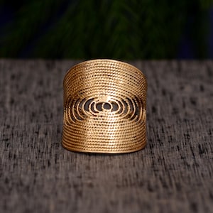 Puede incluir: Un anillo dorado con un diseño tejido y texturizado. El anillo presenta un diseño recortado de forma ovalada en el centro, agregando un elemento decorativo. El anillo está sobre una superficie oscura y texturizada.
