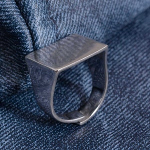 Puede incluir: Un anillo plateado con una parte superior rectangular y una banda gruesa. El anillo está hecho de metal con una superficie ligeramente texturizada. El anillo se muestra sobre un fondo de mezclilla azul.