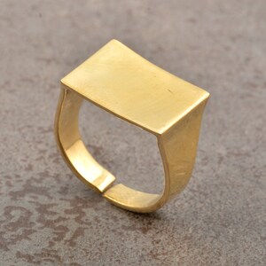 Puede incluir: Un anillo ajustable de color dorado con una parte superior rectangular. El anillo tiene un diseño simple y moderno y está hecho de un material metálico brillante. La banda está abierta en la parte inferior, lo que permite ajustar la talla.