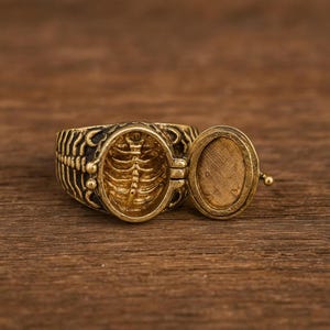 Puede incluir: Un anillo de metal dorado con diseño de relicario con bisagras. La banda exterior muestra un patrón de caja torácica texturizado. Al abrirse, el interior revela un relieve detallado de una caja torácica y una tapa interior ovalada lisa.