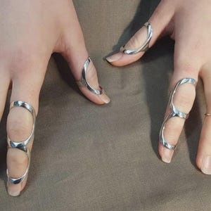 Puede incluir: Una colección de anillos tipo férula para dedos hechos de metal plateado, usados en varios dedos. Los anillos presentan un diseño abierto y curvo que envuelve las articulaciones, proporcionando una apariencia elegante y pulida.