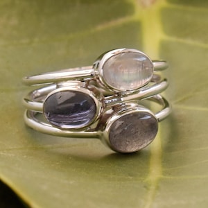 Puede incluir: Tres anillos de plata apilados, cada uno con una piedra preciosa ovalada. Las piedras preciosas son de color claro e iridiscente, morado oscuro y gris. Los anillos están sobre un fondo verde.