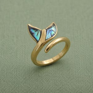 Puede incluir: Un anillo de tono dorado con un diseño de cola de ballena. Las dos secciones de la cola están incrustadas con concha de abulón iridiscente, que muestra tonos azules y verdes brillantes. La banda está abierta, lo que permite un ajuste regulable.