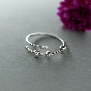 Puede incluir: Un anillo abierto de color plateado que presenta una banda circular delgada con tres pequeñas cuentas esféricas y lisas que se deslizan a lo largo del alambre. El anillo tiene una abertura en la parte superior y los extremos son redondeados.