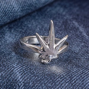 Puede incluir: Un anillo plateado con un diseño de estrella, con un elemento central puntiagudo y múltiples puntas radiantes. Una pequeña gema redonda está engastada en la base de la punta central. El anillo se muestra sobre una superficie de mezclilla azul.